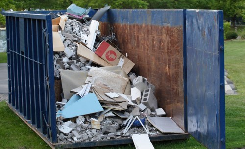 An open-top blue industrial skip filled with mixed construction and household debris, including broken tiles, concrete blocks, pieces of plasterboard, cardboard boxes, and scrap wood, situated outdoors on a grassy area with trees in the background. The skip's interior shows signs of rust and wear on the metal walls, with some loose debris spilling over the edges onto the ground. The scene indicates rubbish collection and removal services, typical of commercial waste management in Aldershot or surrounding areas, provided by a specialised waste disposal company. The natural lighting highlights the textures of the materials, demonstrating the variety of waste types handled by Commercial Waste Aldershot, aligned with their rubbish removal offerings in the local postcode region.