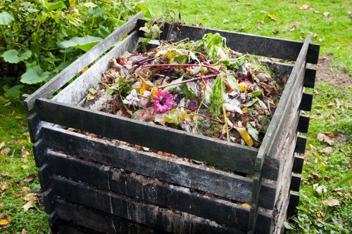 A black, wooden compost bin filled with organic waste materials such as plant trimmings, fallen leaves, and flower debris, located outdoors on a grassy area with green foliage in the background. The waste appears to include a mix of green and brown materials, some of which are slightly decomposed. The bin is situated on a garden or yard surface, and the image suggests a setting suitable for waste collection and composting services offered by Commercial Waste Aldershot, part of their rubbish removal operations in the local area near Aldershot. The natural lighting highlights the textures of the organic matter and the weathered surface of the wooden bin.