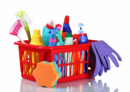 A red plastic basket filled with various cleaning and household products, including spray bottles, scrub brushes, sponges, and rubber gloves, arranged on a white surface with a plain background. In the foreground, a multi-coloured sponge with a rainbow pattern and rounded edges leans against the basket. The products are made of plastic and rubber, featuring bright colours such as yellow, blue, red, green, and purple, with some bottles having clear or white sections. The rubber gloves drape over the right side of the basket, while other cleaning tools and bottles are visible both inside and slightly on top of the basket, suggesting a recent or ongoing cleaning activity. The overall scene depicts typical household cleaning supplies, with textures ranging from smooth plastic to soft rubber, and a clean, well-organized presentation. This image can be associated with rubbish and waste management services provided by Commercial Waste Aldershot, which include collection and disposal of household cleaning waste from domestic environments in the Aldershot area, within the postal code region inferred from the context.