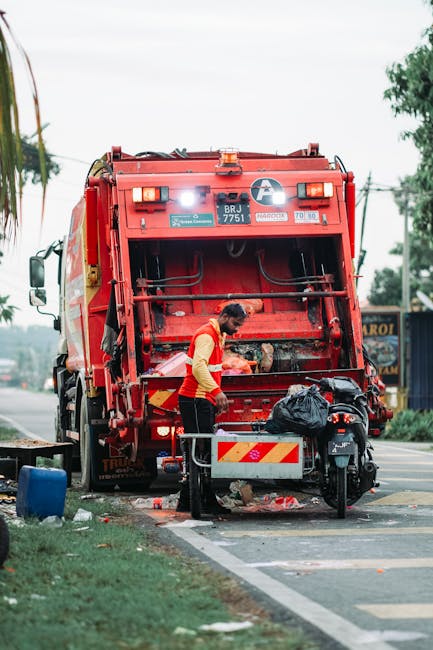 A middle-aged man with dark hair is seen disposing of waste into a stainless steel public trash bin located on a paved outdoor walkway. The man is wearing a black T-shirt with a partially visible grap
